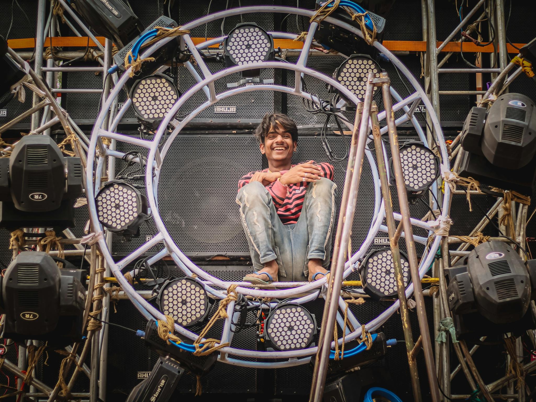 Reviews A man joyfully sitting on a stage lighting rig in Ballia, India, showcasing festival preparation.